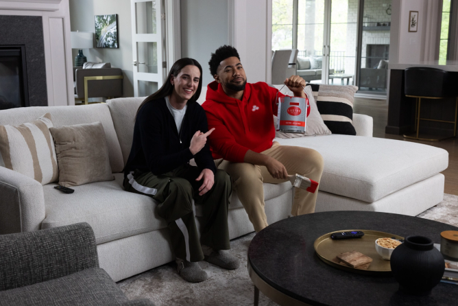 Caitlin Clark and Jake from State Farm pose on couch with bucket of paint for State Farm "From the Logo" ad.