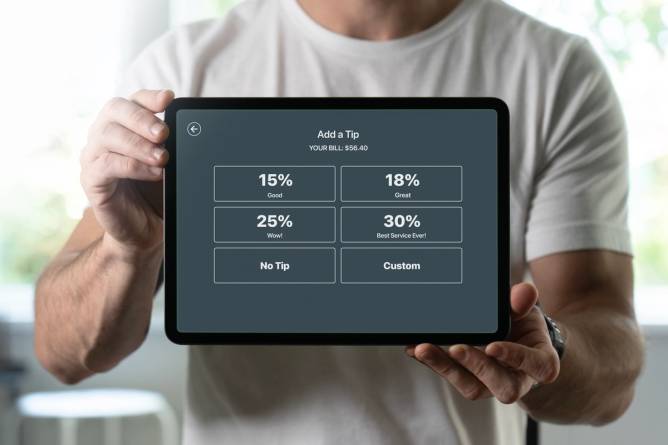 A man holds a tablet with tipping options from 15% to 30%. 