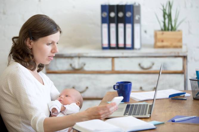 A woman works in front of her laptop while holding a baby in one hand and a phone in the other.