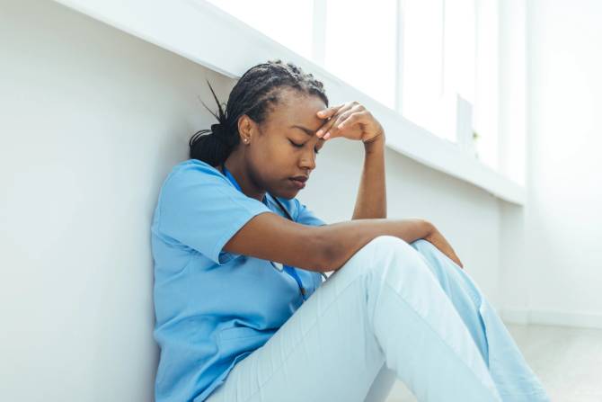 A tired Black nurse sits on the floor with her hand on her head.