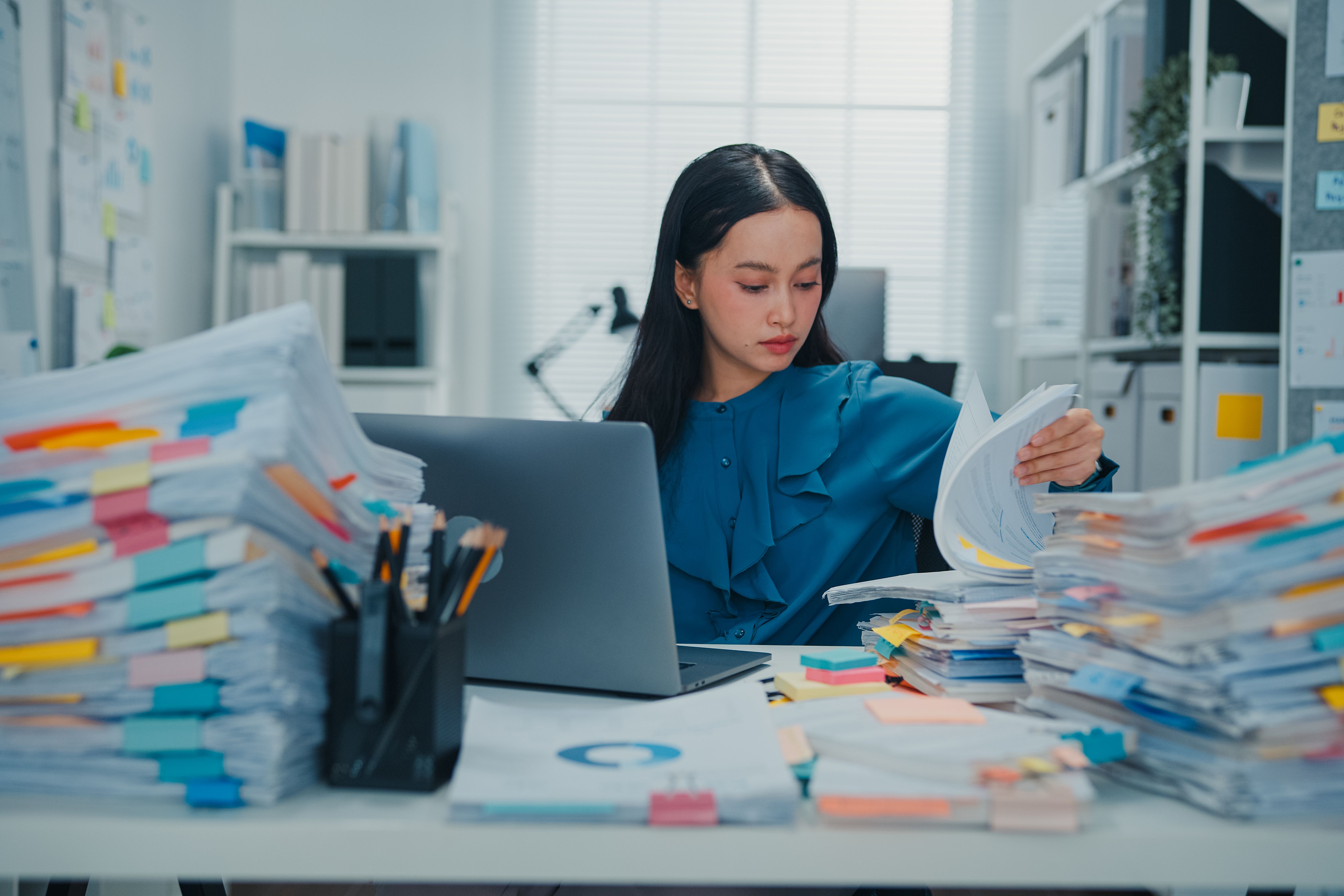 Woman sitting at desk looking through stacks of reports with laptop.