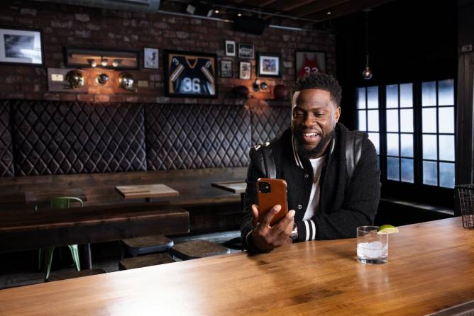 comedian Kevin Hart looks at his phone inside of a dark bar with a drink in front of him in an image from a new ad promoting DraftKings 