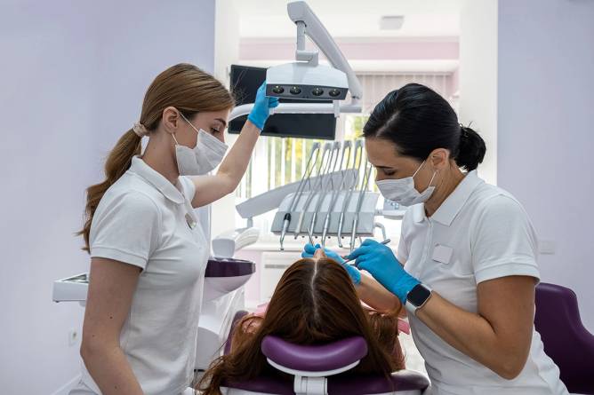 Female dentist and assistant treat the teeth of a female patient.