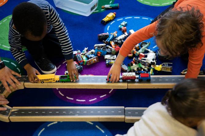 A group of children enrolled in a Head Start childcare program play with a wooden train and vehicle toy set.