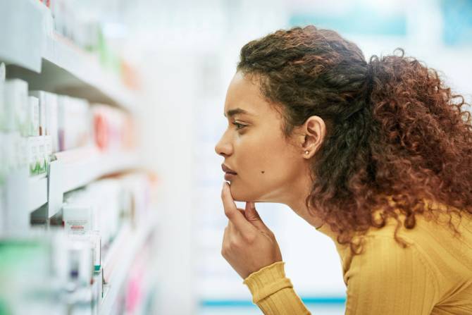 A woman with tight brown curls and a yellow sweater ponders in front of a pharmacy shelf.