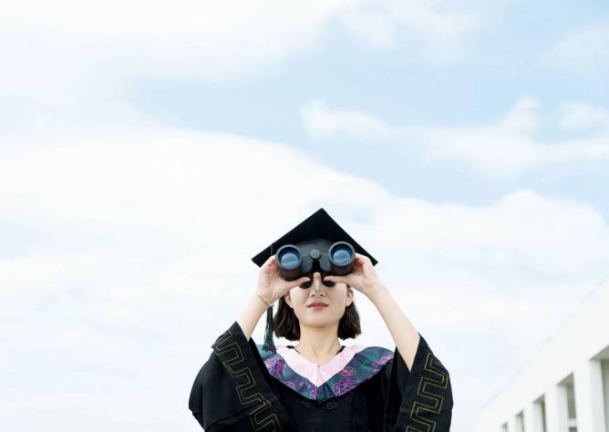 A young woman wearing a graduation cap and gown looks into the distance using binoculars.