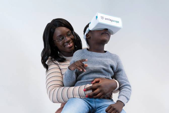 A woman smiles as she holds her child, who is wearing a VR headset