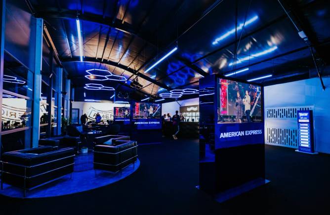 a photo of a dimly lit blue-hued lounge with black leather chairs, a bar, and American Express signage that is part of American Express's F1 activation at the Las Vegas Grand Prix