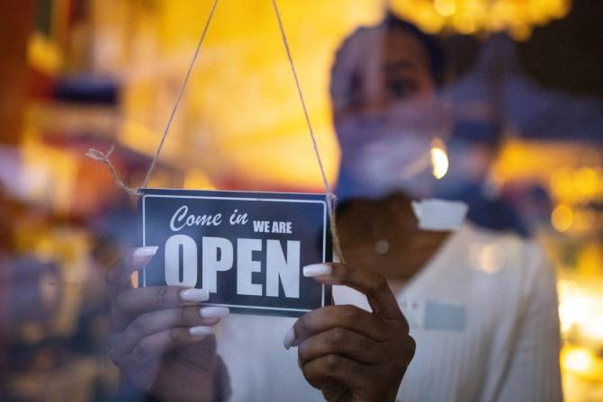 A woman turning a "Come on in we are open" sign behind a glass window.