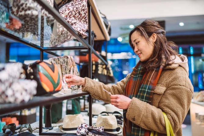 A woman checks the price of a handbag on a store shelf.