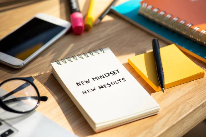 a desk with an iPhone, highlighter, pen, eyeglasses, and a notebook that says new mindset new results