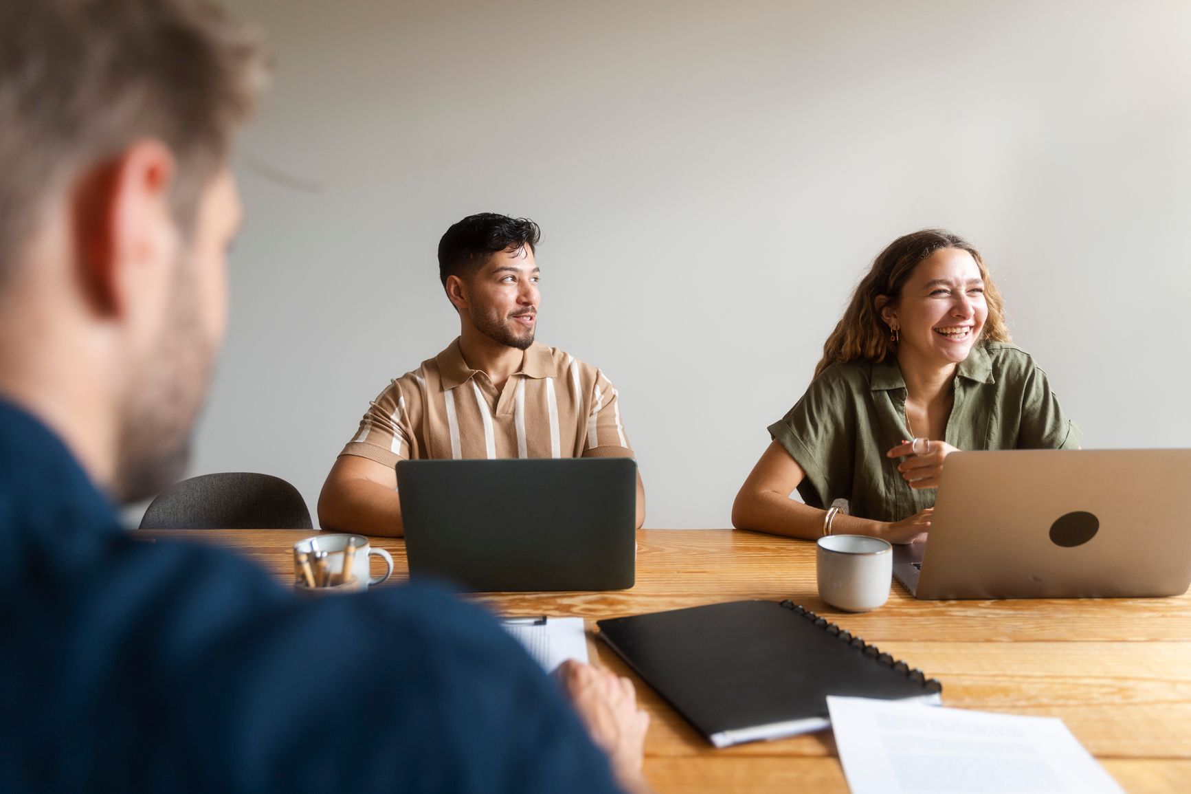 Three colleagues laugh and chat during a casual office meeting around a wooden table, with open laptops and coffee mugs in front of them.