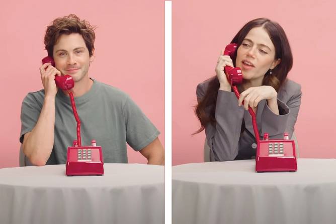 Two people at a table covered with a white tablecloth, they're both holding the receiver of a red corded phone.