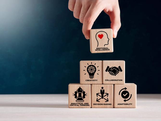 A hand puts a block that reads "emotional intelligence" on top of a pyramid of wooden building blocks. 