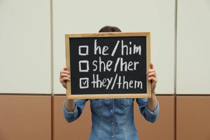 nonbinary person holding up a chalkboard with he/him she/her they/them next to check marks with they them checked