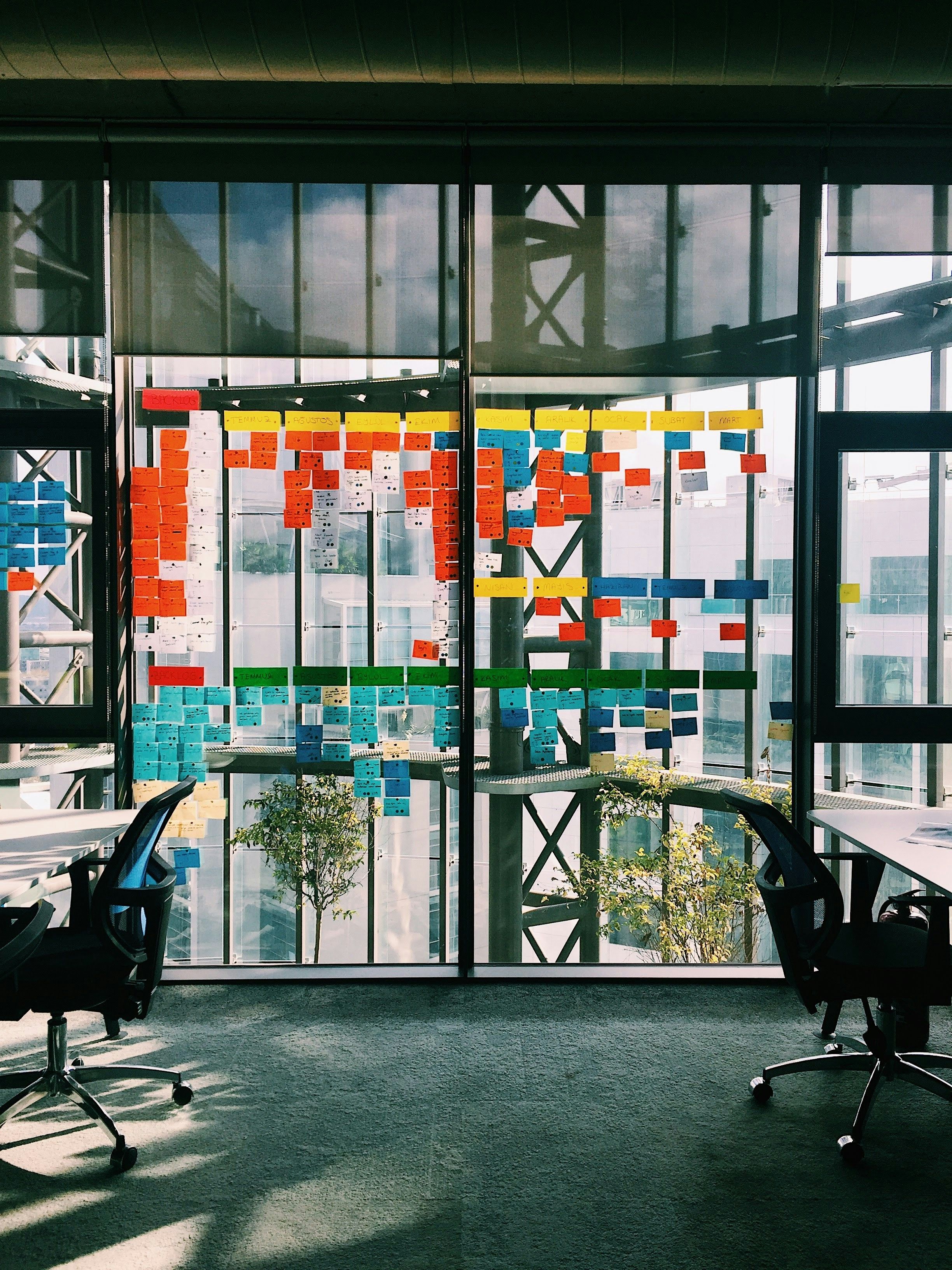 A floor-to-ceiling glass office window covered in hundreds of colorful sticky notes arranged in a grid pattern, with two empty office chairs and desks on either side.