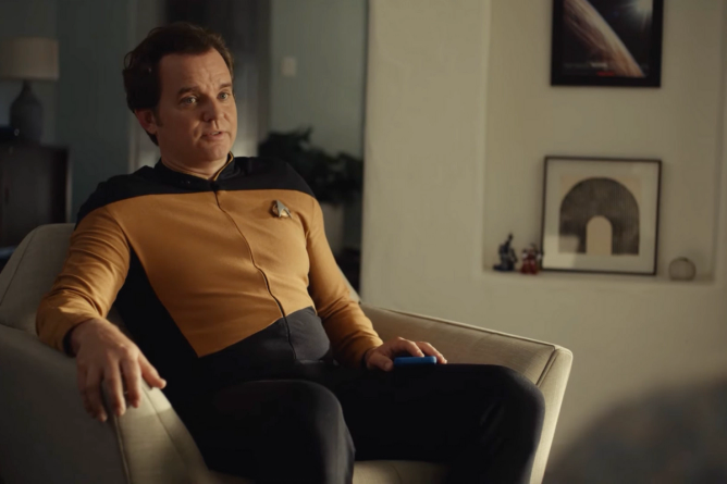 An actor wearing a Star Trek uniform and portraying a programmer for Pluto TV sits in a living room with framed art in the background