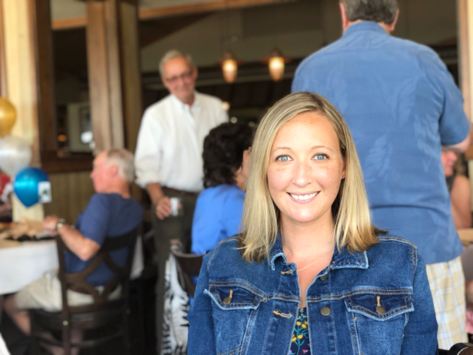 A smiling woman with short blonde hair and a jean jacket, looking straight to camera.