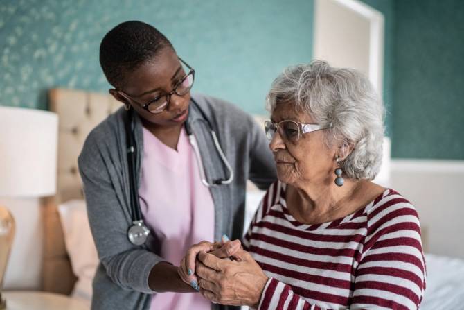 A caregiver in pink scrubs and grey sweater helps an elderly woman stand up.