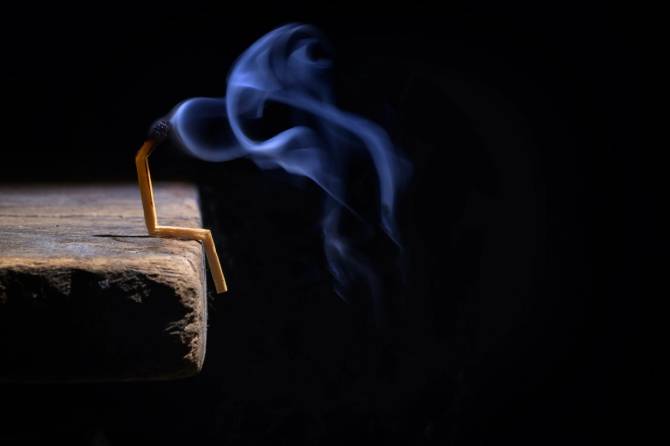 a burned out match, bent in a way that resembles a person, sitting on the edge of a plank of wood, with a black background