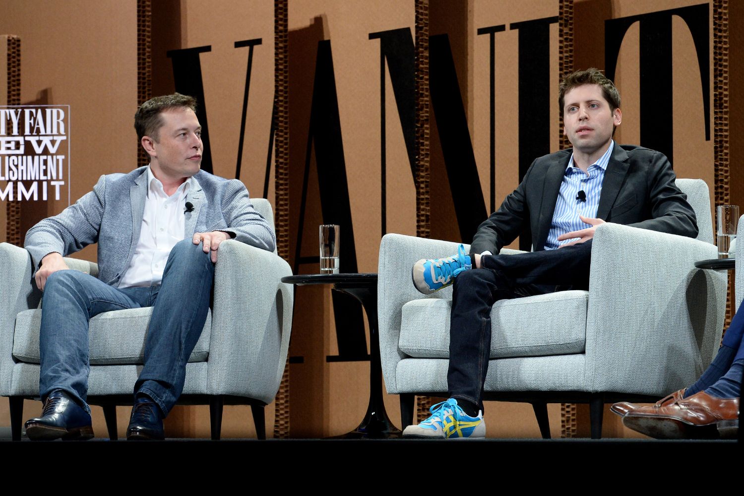 Elon Musk and Sam Altman sit in gray armchairs next to each other on a stage.