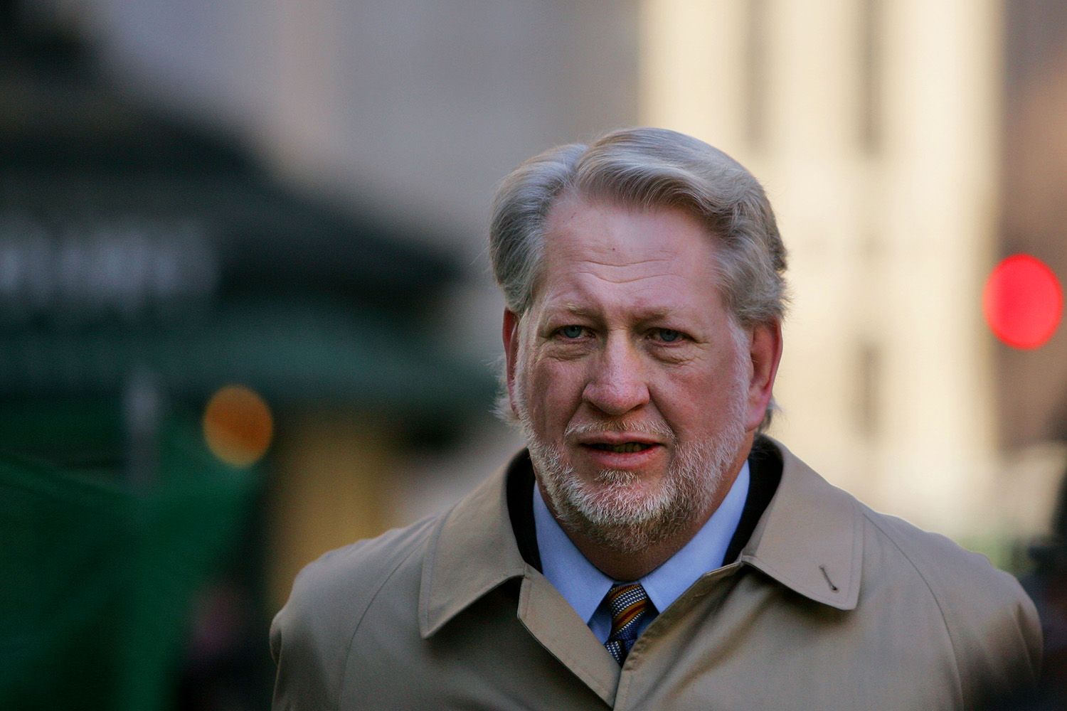 A middle-aged man with greying hair wears a trench coat and a frustrated look on his face.