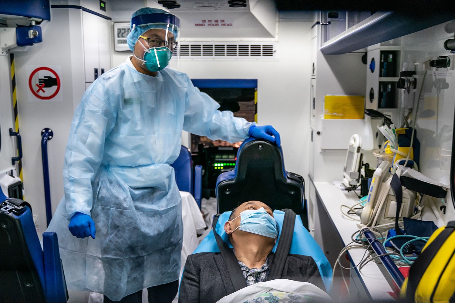 A healthcare worker wearing a mask, face shield, disposable lab coat and gloves stands over a person on a gurney, also wearing a mask
