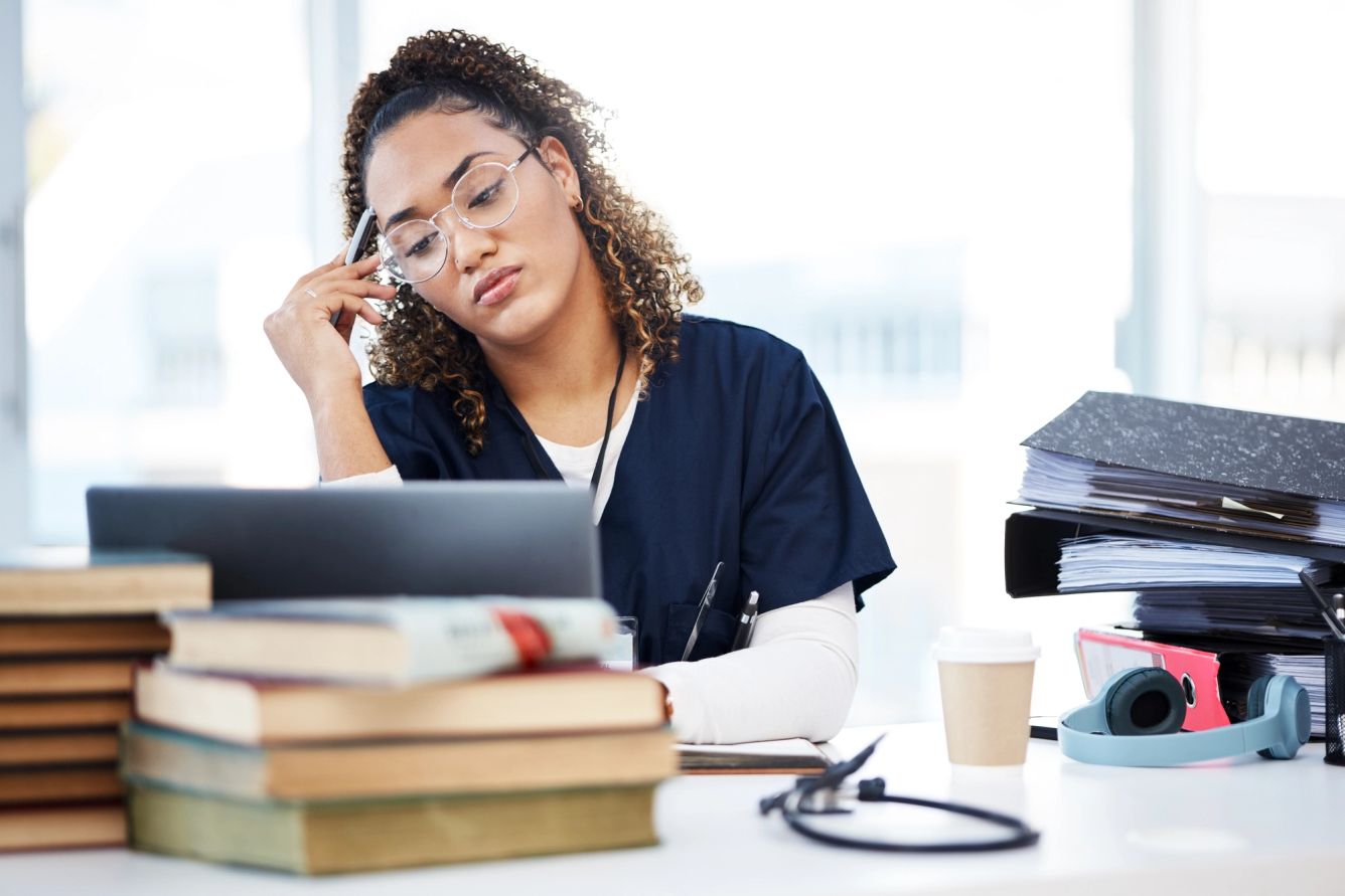 Nurse looking stressed while looking at laptop with books around her and a coffee cup nearby