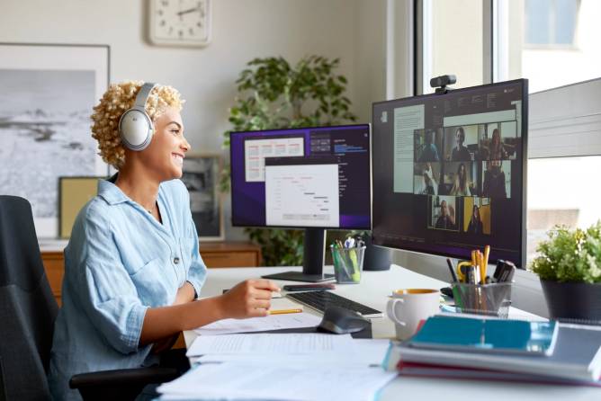 A woman sits at a desk in front of a computer monitor, taking a video call.
