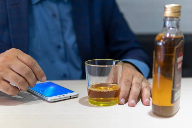 A person holds an alcoholic drink in one hand while scrolling on their phone with another.