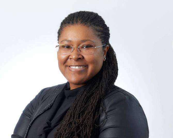 A Black woman with locs and glasses smiles at the mirror.
