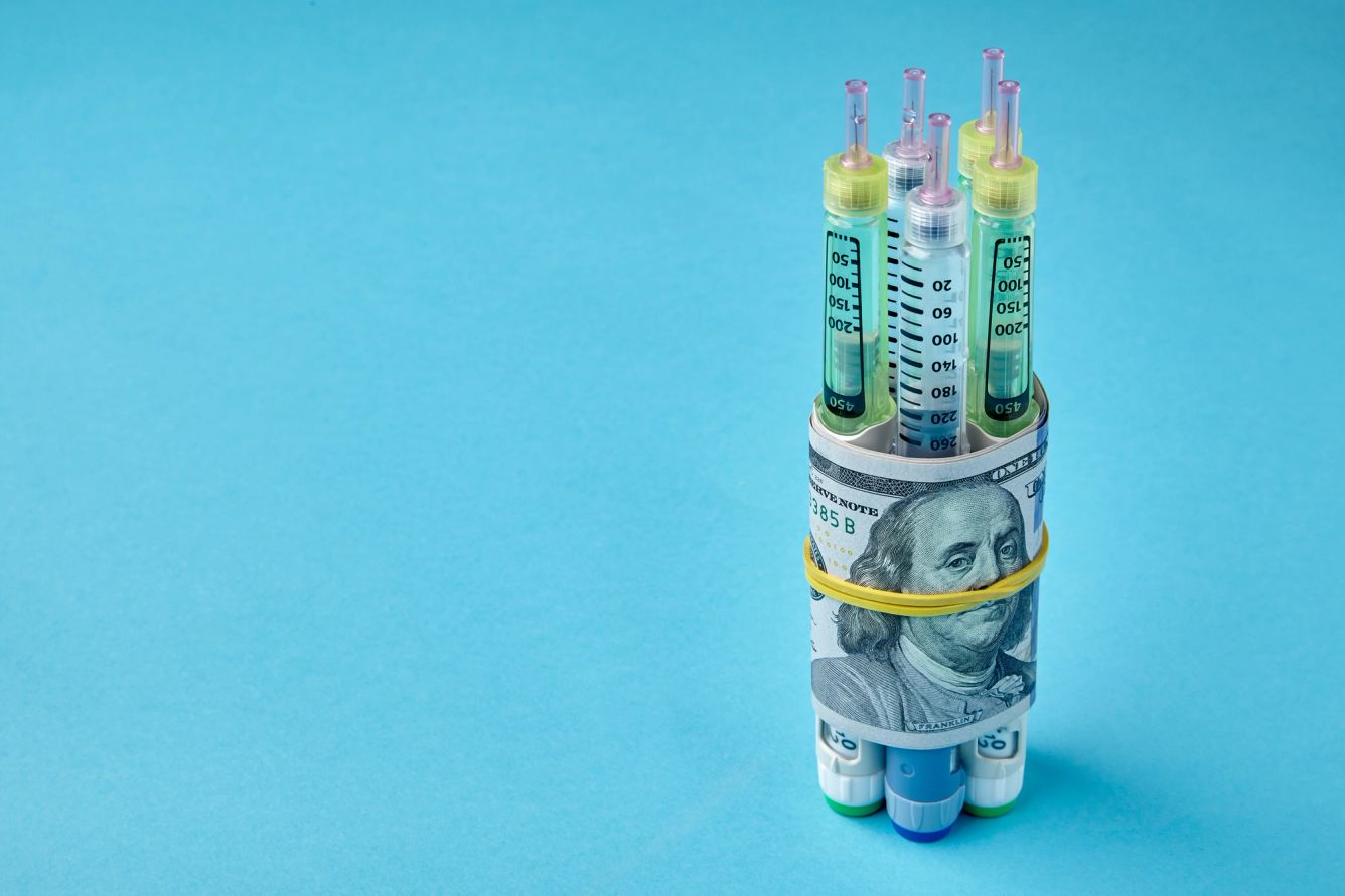 Insulin syringe pens wrapped in dollar bills on a blue background.