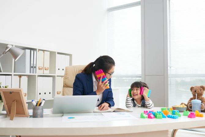 A woman sits at a desk next to a young girl playing with legos.