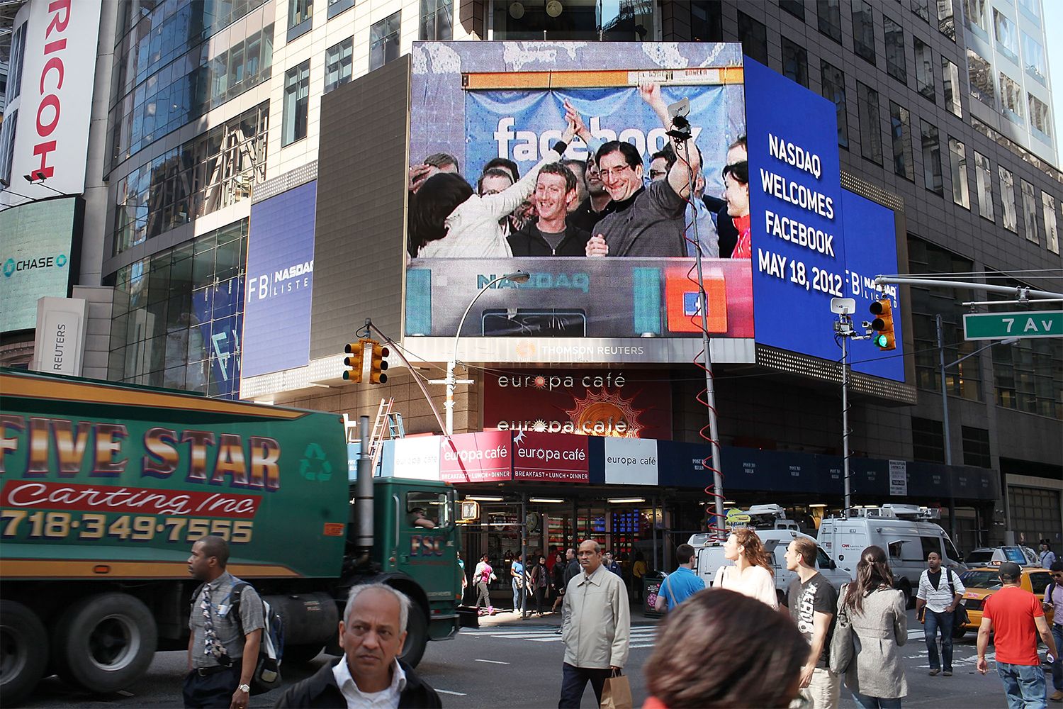 A large screen on the side of a building shows Mark Zuckerburg at the NASDAQ podium at the Stock Exchange, text reads, 