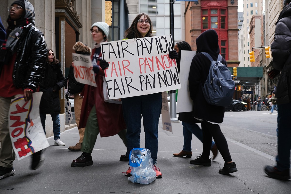 A woman holds a sign that reads Fair Pay Now in the middle of a picket line.