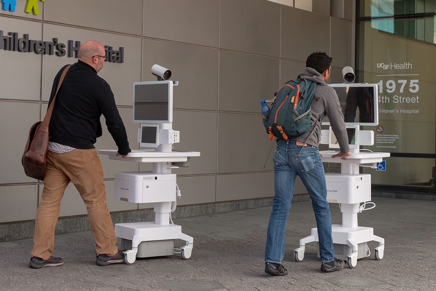 Two staff members wheel telemedicine carts into the entrance of a hospital building.