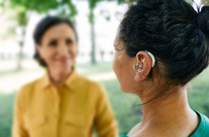 A woman uses a hearing aid to communicate with her female friend in a park.