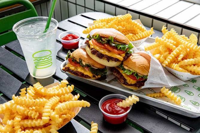 burgers, fries, and a drink in a branded Shake Shack cup on a picnic table with a green chair