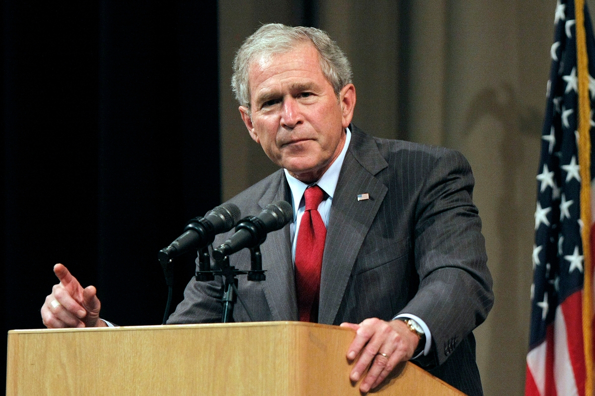 President George W. Bush standing at a podium, one arm raised, open palm, speaking.
