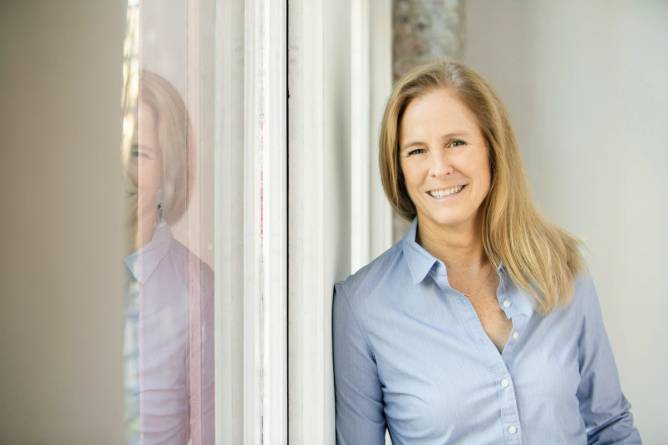 A smiling blonde older woman leans against a mirrored surface