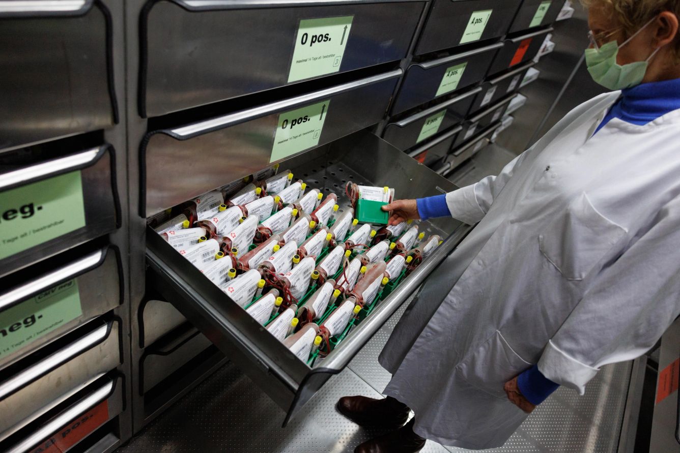 Masked doctor holding a bag of blood and opening a drawer filled with more blood bags in a room of drawers