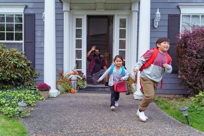 Two kids running outside the house while a dad waves by the door.