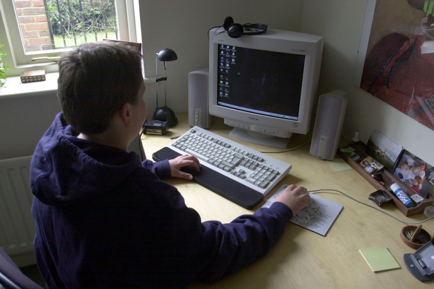 A teenager in a hoodie sits at a desk interacting with a boxy desktop computer, wired mouse, and keyboard.