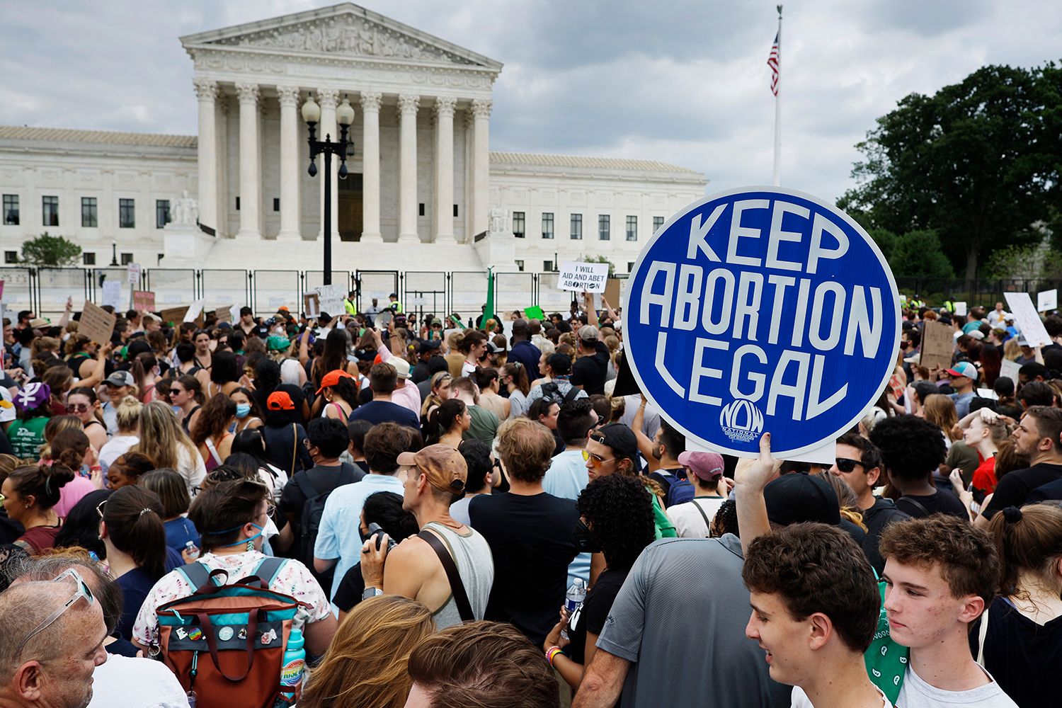 A large crowd of protesters stand in front of the Supreme Court, a young man holds a sign above his head in the shape of circle that reads, 'Keep Abortion legal'