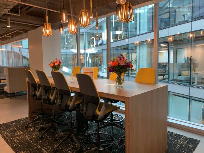 A conference table with several chairs beneath warm lighting and floor to ceiling windows behind it