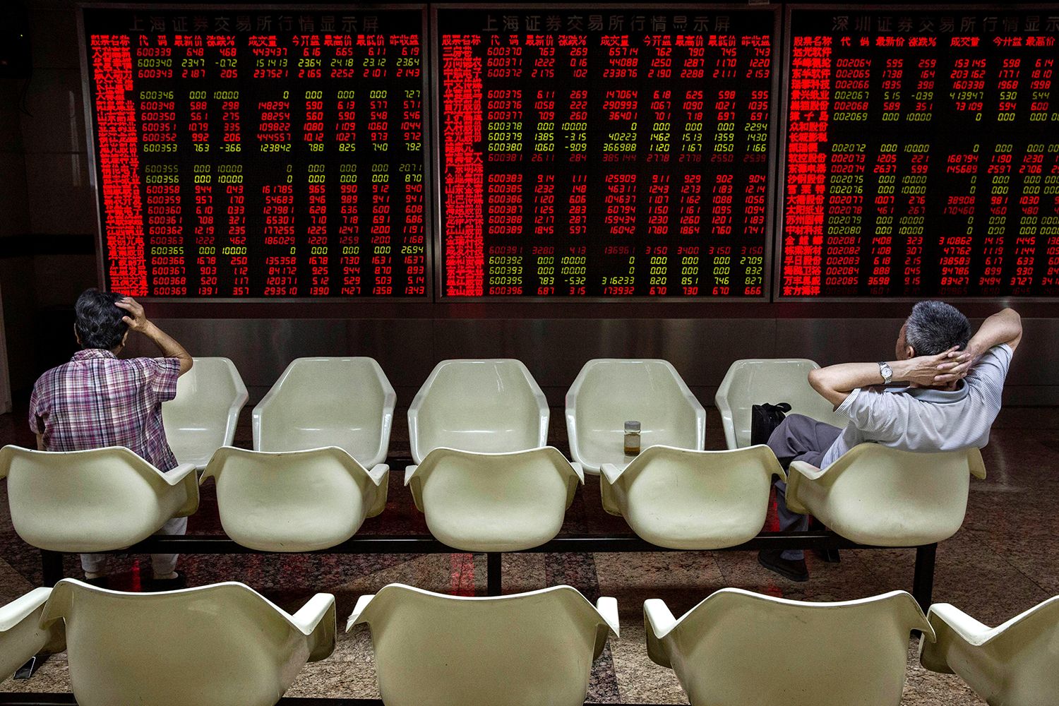 Two men sit in chairs in front of a wall of stock tickers at a local brokerage house.
