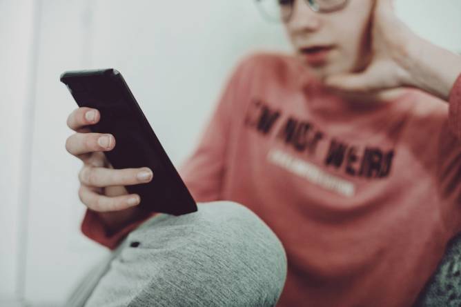 A teen with glasses leans on a couch using a phone.