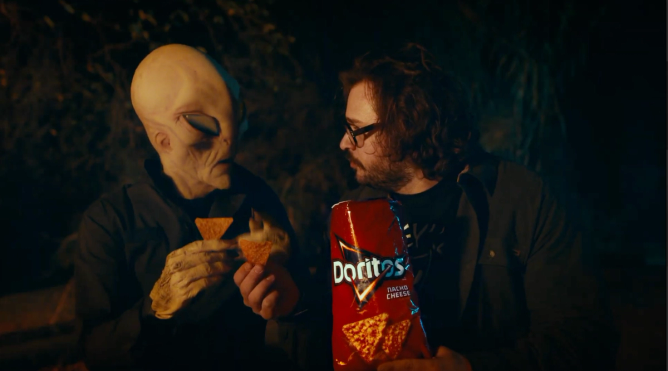 A person dressed as an alien holds a chip and sits next to a man in glasses, who is holding a red Doritos bag. 