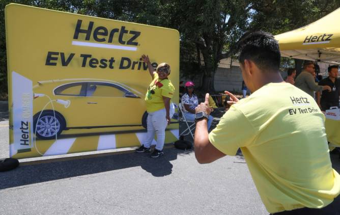 A woman has her picture taken in front of a yellow Hertz sign that says "EV test drive."