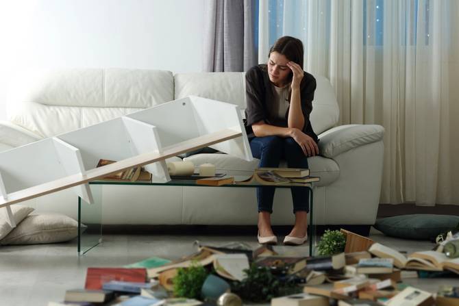 A sad woman sits on her couch surrounded by a destroyed apartment. 